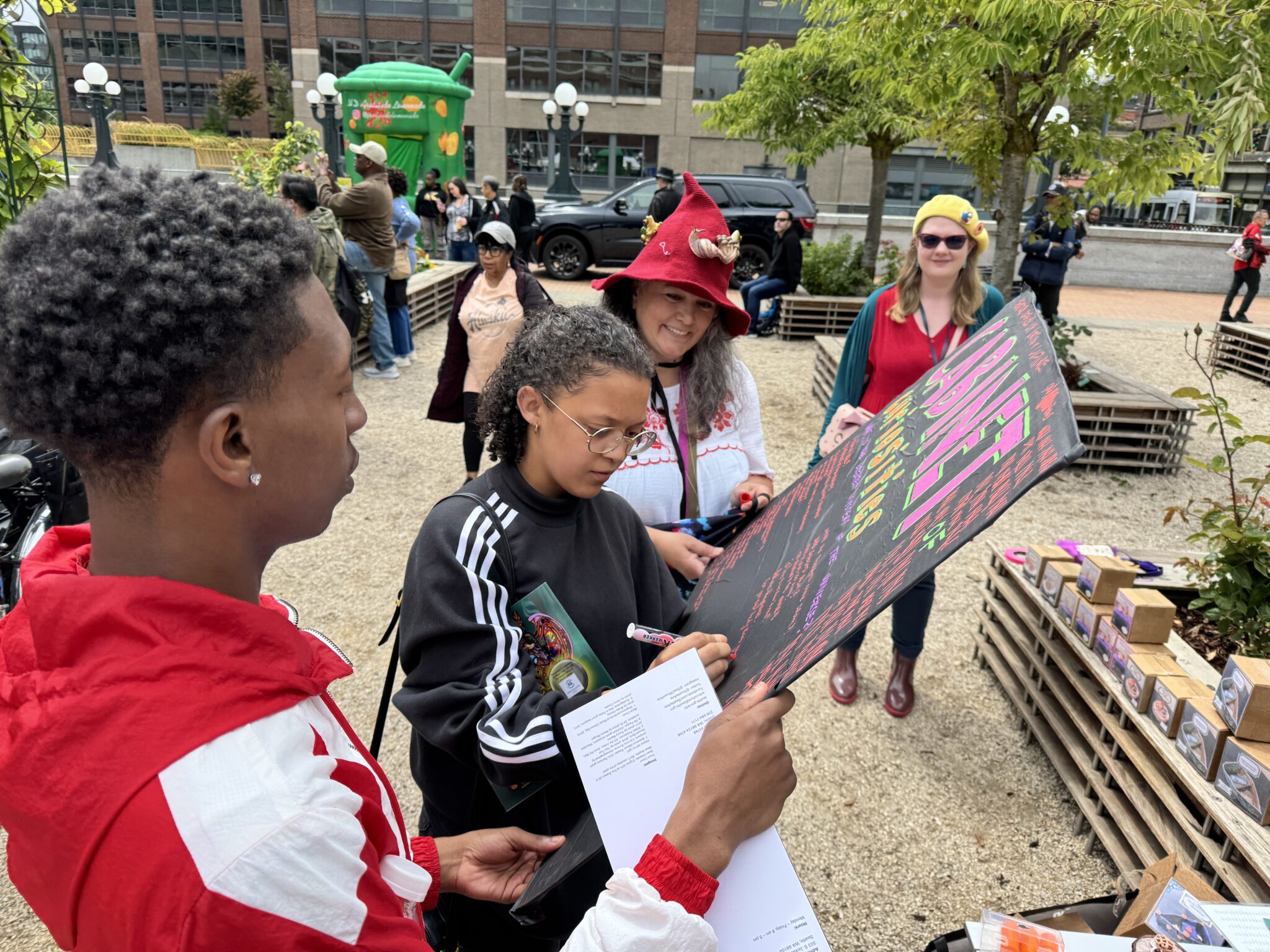 A person in a red and white track suit holds up a sign that a person with a curly ponytail and glasses writes on.