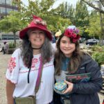 Artist Angie Hinojos smiles with her arm around another person with long hair that holds a small box of gifted art. Angie wears a red felted pointy hat adorned with flowers and a white shirt with floral embroidery, the person next to her wears a flower crown atop their head.