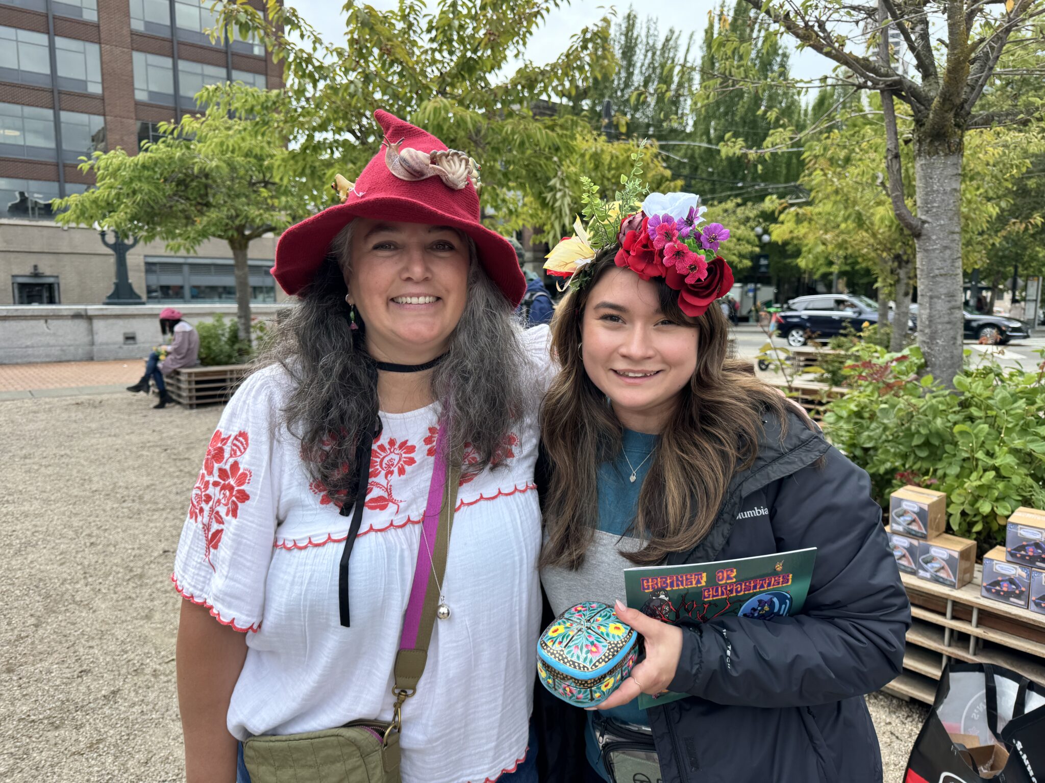 Artist Angie Hinojos smiles with her arm around another person with long hair that holds a small box of gifted art. Angie wears a red felted pointy hat adorned with flowers and a white shirt with floral embroidery, the person next to her wears a flower crown atop their head.