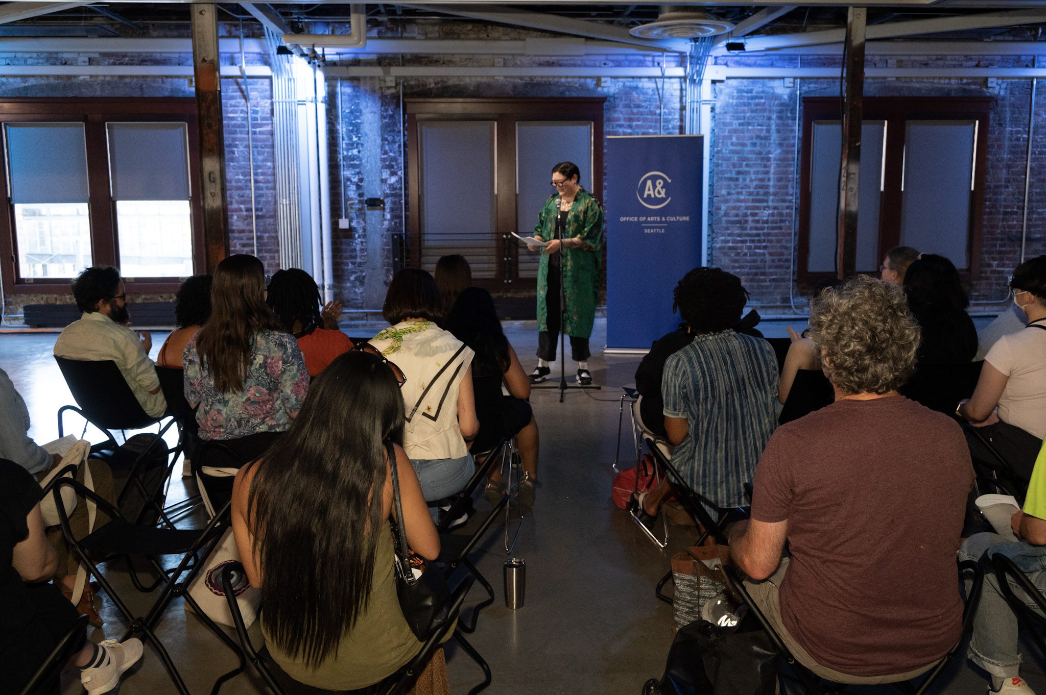 A crowd of people sit watching a person with short hair, glasses, and a long green coat speak into a mic.