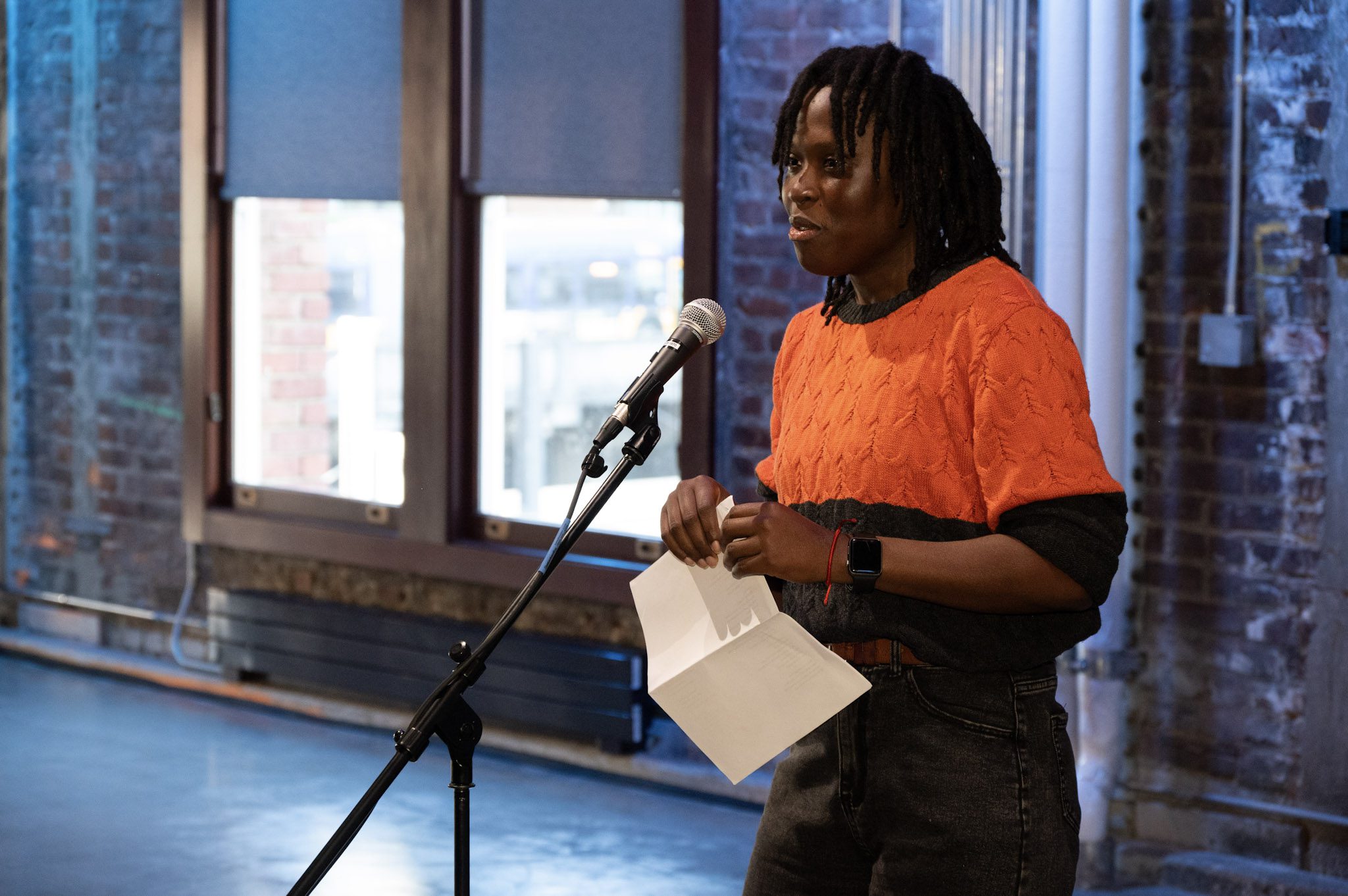 A dark-skinned person with twists and a bright orange sweater speaks into a microphone.