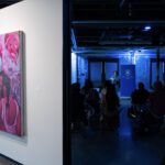A Painting in purple tones hangs on a wall to the left and a crowd of people lit in blue lighting sits to the right.