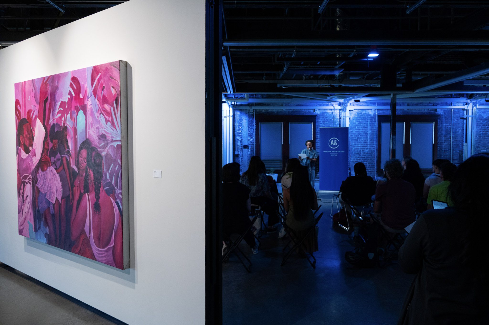 A Painting in purple tones hangs on a wall to the left and a crowd of people lit in blue lighting sits to the right.
