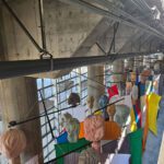 The Guests from above and behind shows the back of their heads, fabric colors, and armatures that they hang from. The space has fencing, brick walls, and the underside of Overlook Walk.