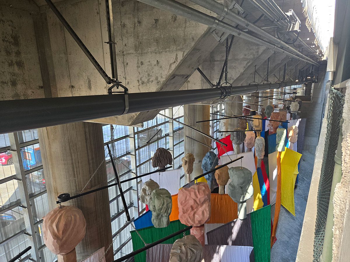The Guests from above and behind shows the back of their heads, fabric colors, and armatures that they hang from. The space has fencing, brick walls, and the underside of Overlook Walk.