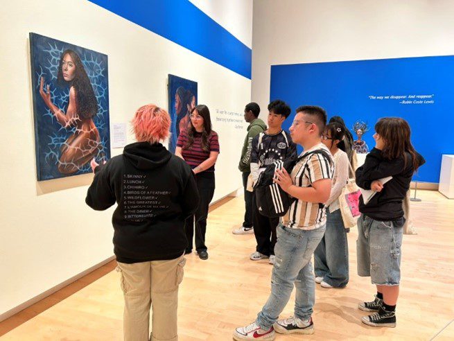 A group of people stands in an art gallery observing a painting of a woman kneeling in water with a net-like pattern around her. One person with pink hair appears to be leading a discussion about the artwork.
