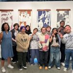 A diverse group of twelve people poses in front of four vertical paper cut-out artworks displayed in a gallery. They are smiling and making playful gestures, standing on a polished concrete floor in a well-lit space.