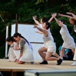 Six figures in white dance on an outdoor stage.