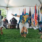 Three Indigenous dancers in traditional dress perform under a large tent.