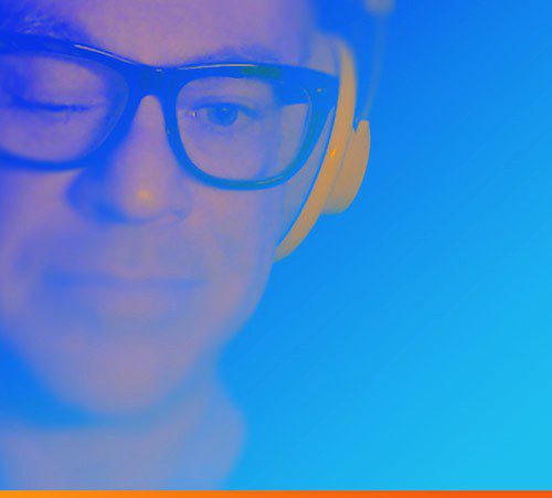 A close up shot of a man wearing glasses and headphones with one eye closed in tones of blue.
