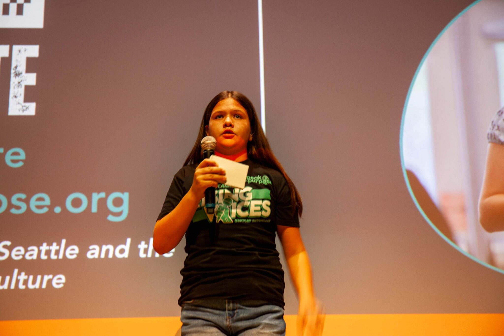 Sam has long dark hair and stands on stage holding a mic