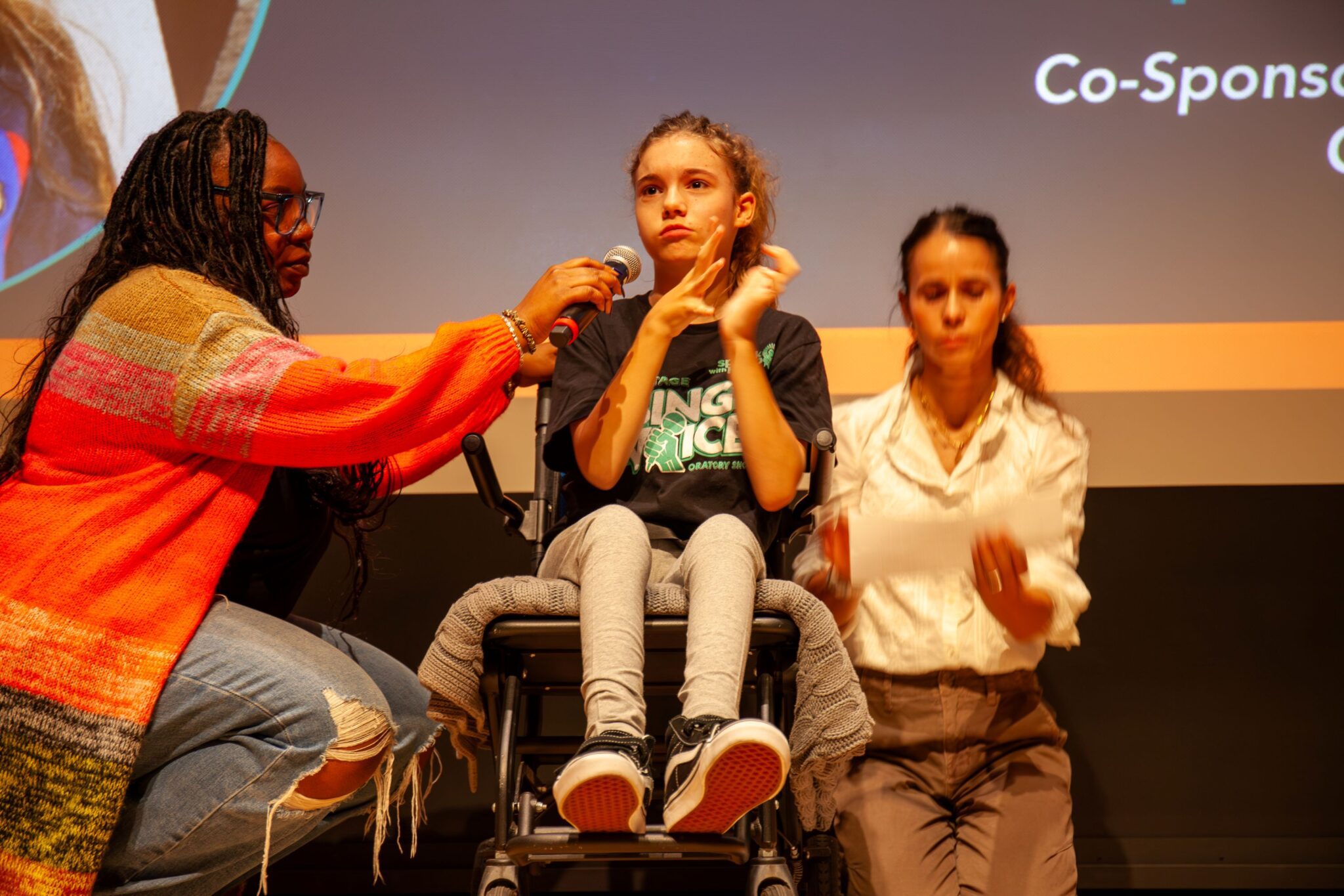 Shea sits in a wheelchair on stage while someone hold up a mic for her and another person signs in the back