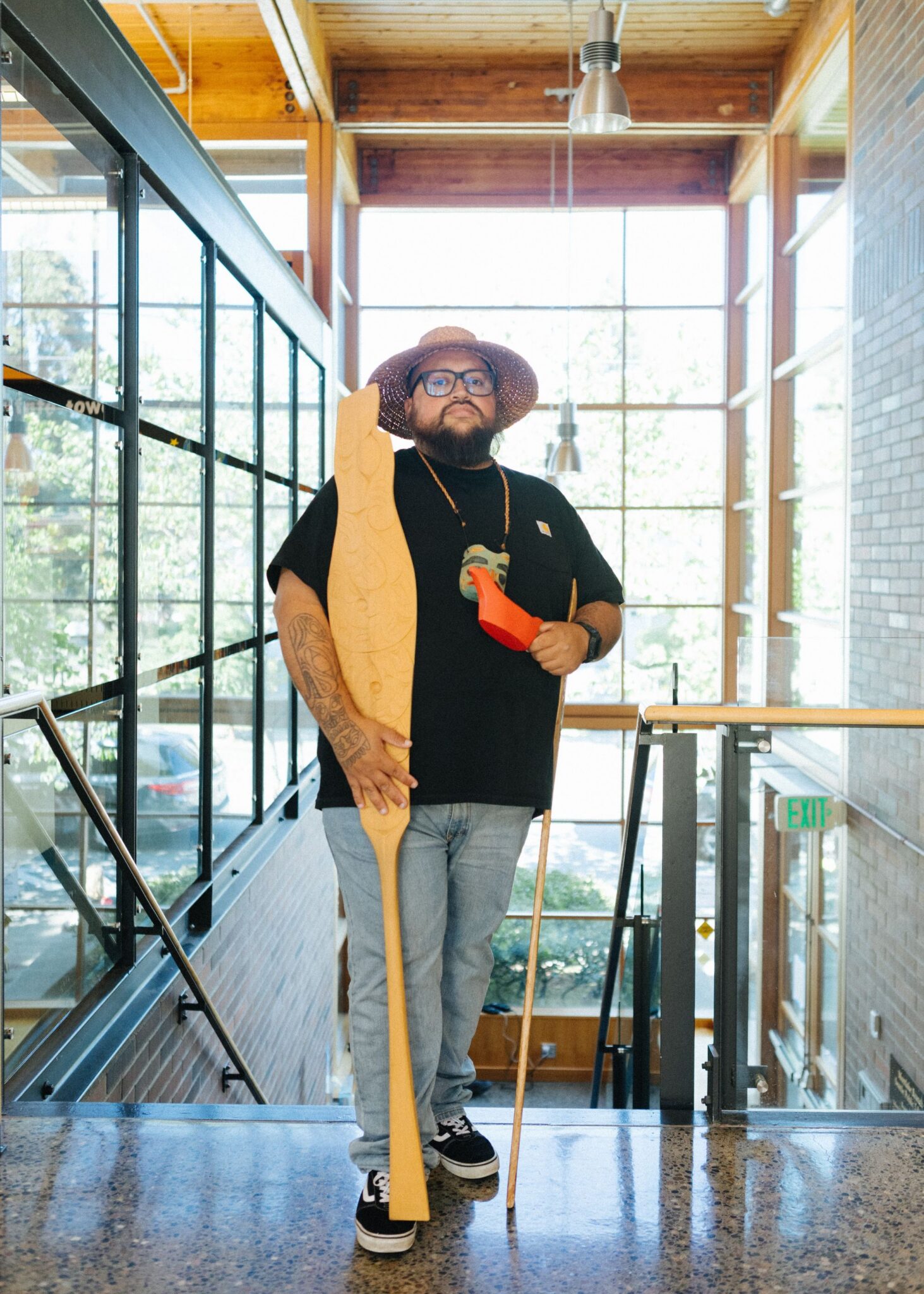 Ty Juvinel standing in a well-lit, modern building. He's holding a long, carved wooden canoe paddle with a detailed, salmon-like design.