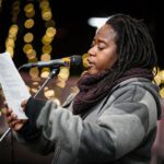 A Black woman with long dreadlocks reads at a mic.