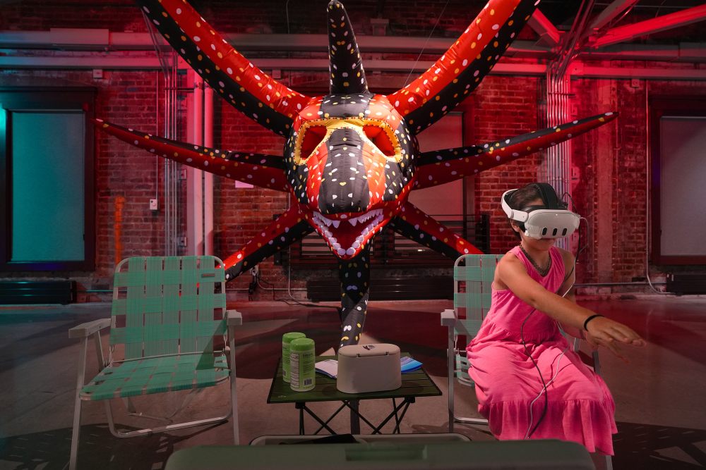 A large inflatable mask sits above two lawn chairs. A girl sits and wears a VR mask, with a hand outstretched.