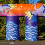 A large, two-legged air vent painted in shades of blue, purple, pink and orange to reflect water and sky, as well two large grey birds with yellow tipped wings