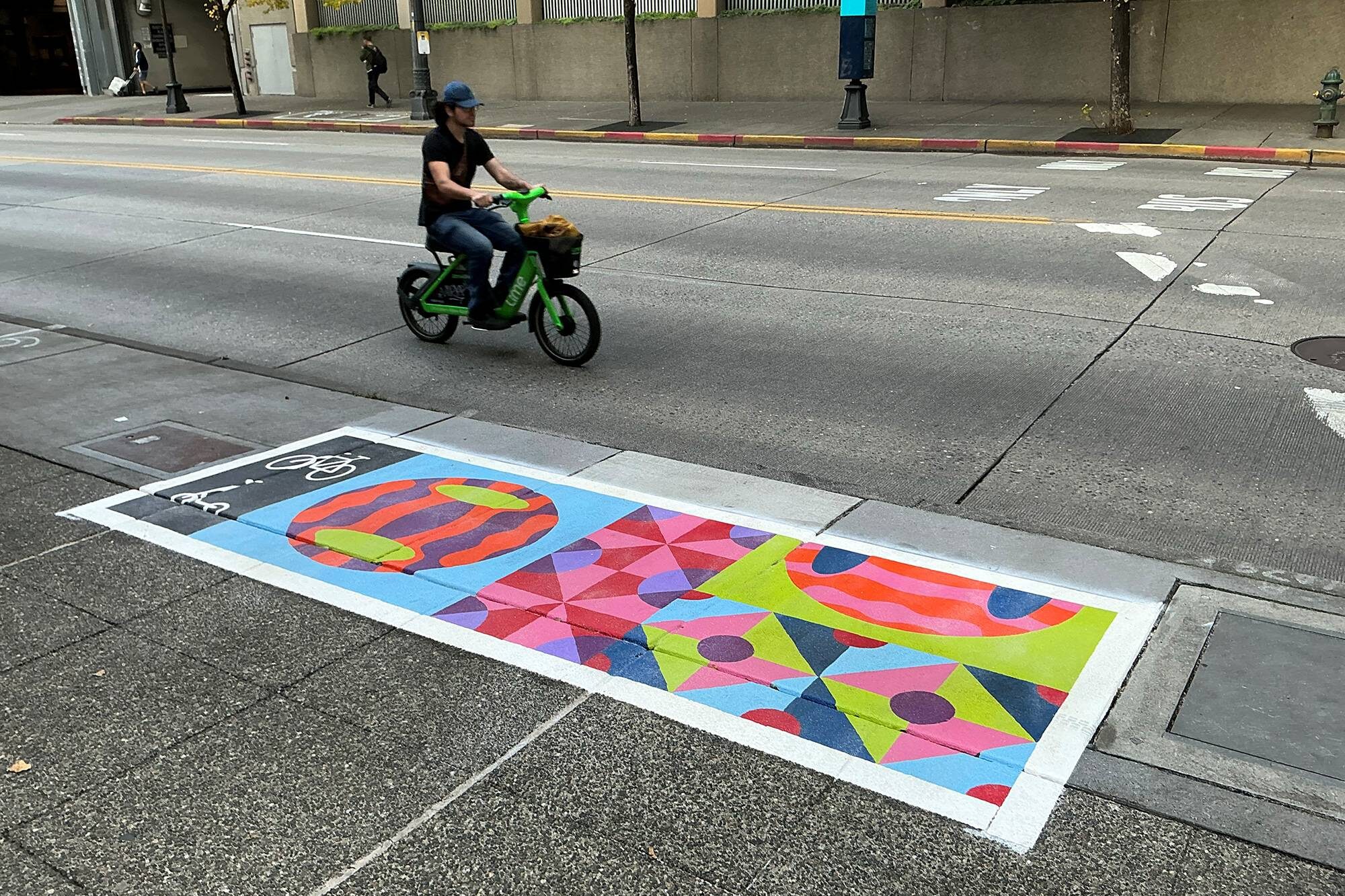 A green bike drives by a large, colorful, abstract painted rectangle on the sidewalk