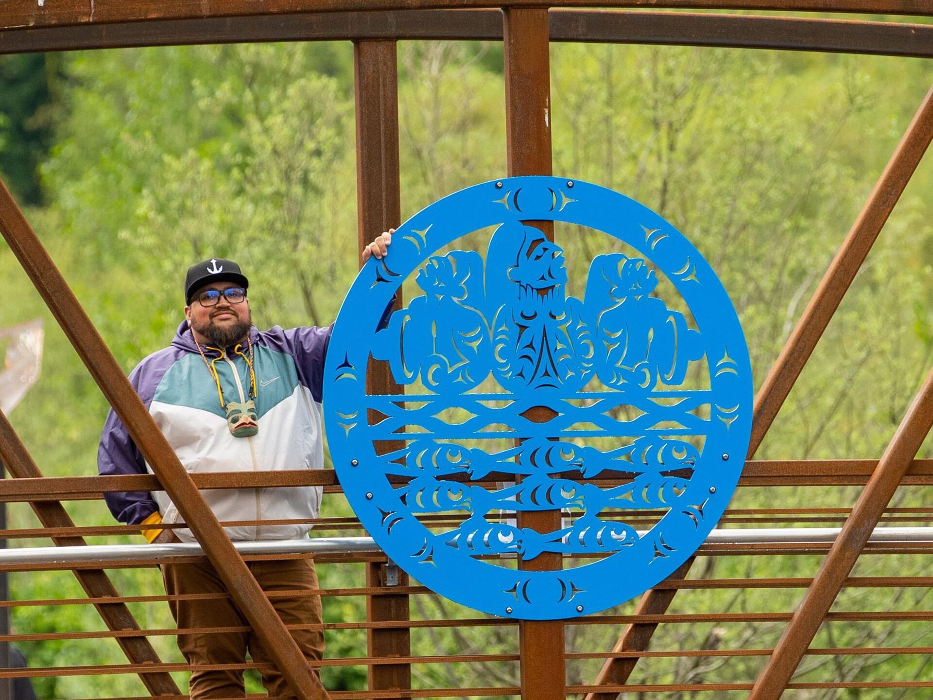 A man wearing a baseball hat standing next to a large circular blue cut out that is half the height of the person and attached to a bridge.