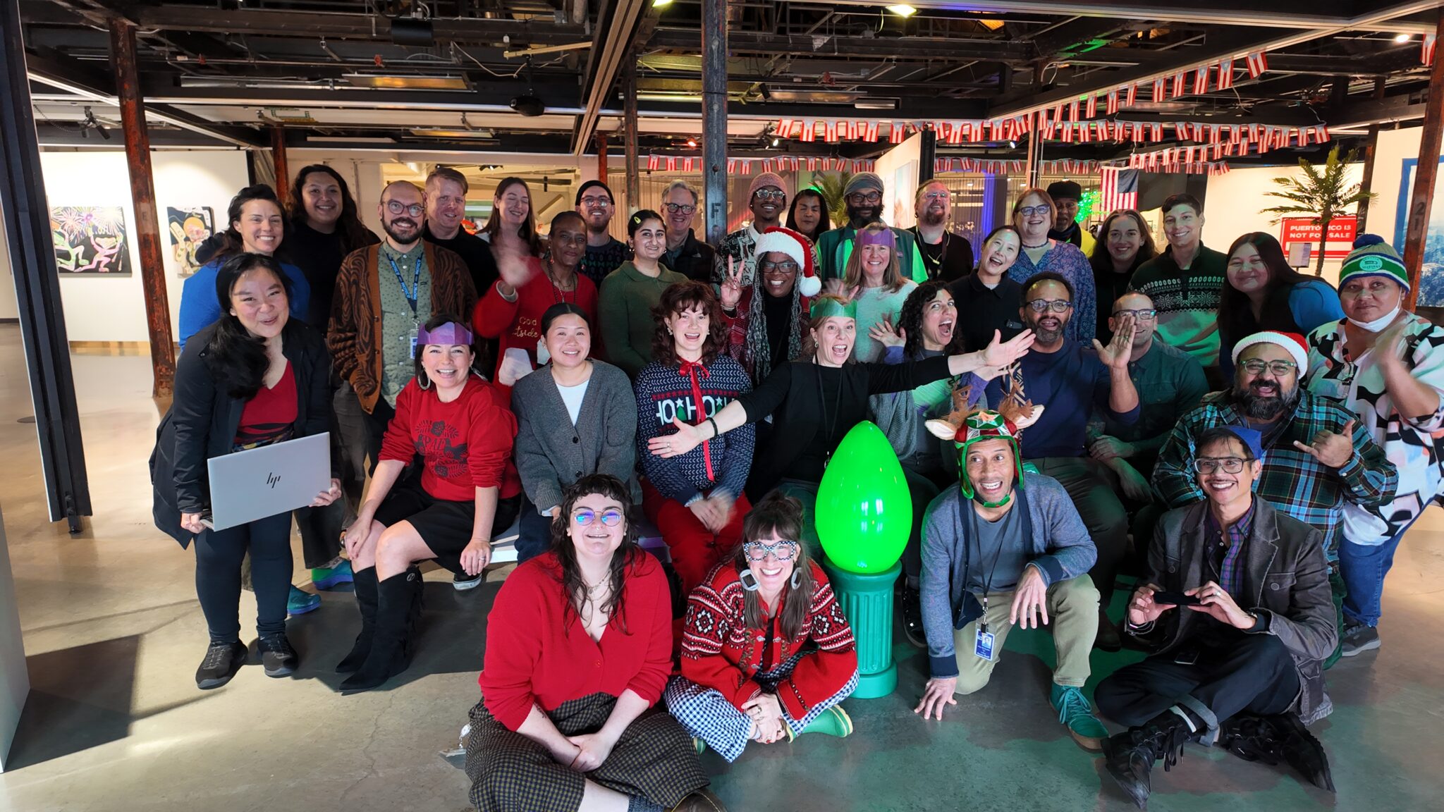 A large group photo of Office of Arts & Culture staff members dressed in holiday outfits with a very large green bulb in the middle