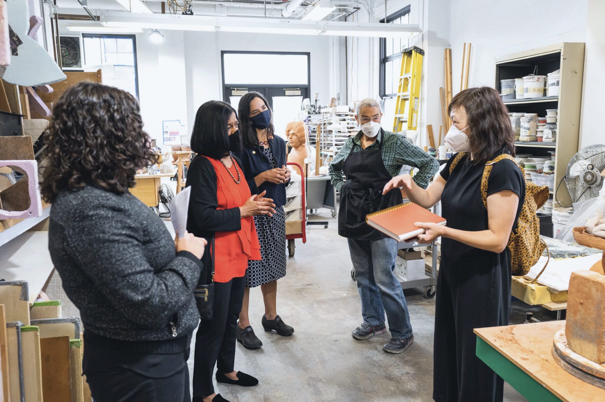 A woman wearing a mask gives a tour to four other women in a large room.