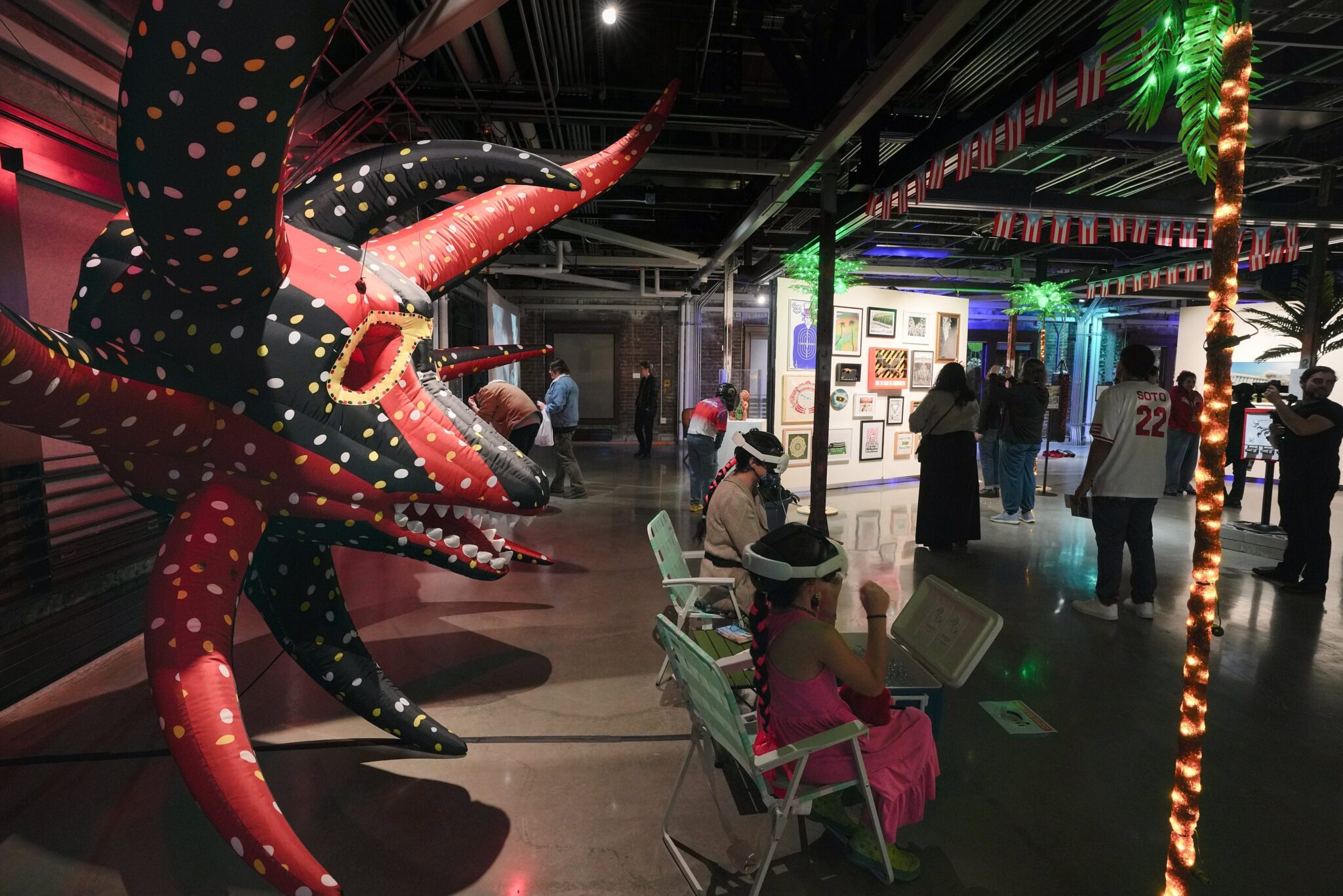 A wide shot of a filled gallery space. A large inflatable red and black mask takes up the left portion of the space with two people sitting wearing VR headsets while others roam the gallery looking at art on the walls.