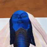 Stylized portrait of a woman from the shoulders up in a white dress and headdress, her skin and hair are blue and her neck is festooned with many necklaces. Her eyes are closed and she is facing up. The horizon is a light blue and the sky is the color of clay.