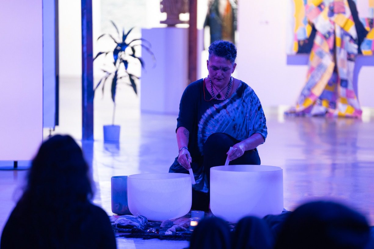 An older woman with short hair sits on the ground in the ARTS at King Street Station gallery and plays sound bowls.