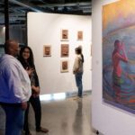 Two people chat in front of a large colorful painting of a figure emerging from the water.