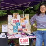 A young adult stands and smiles next to a vendor table set up with many books and zines.