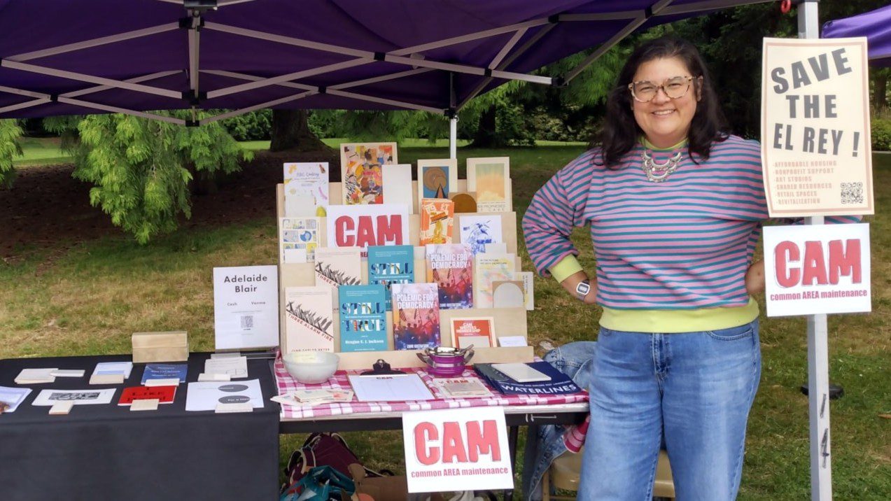 A young adult stands and smiles next to a vendor table set up with many books and zines.
