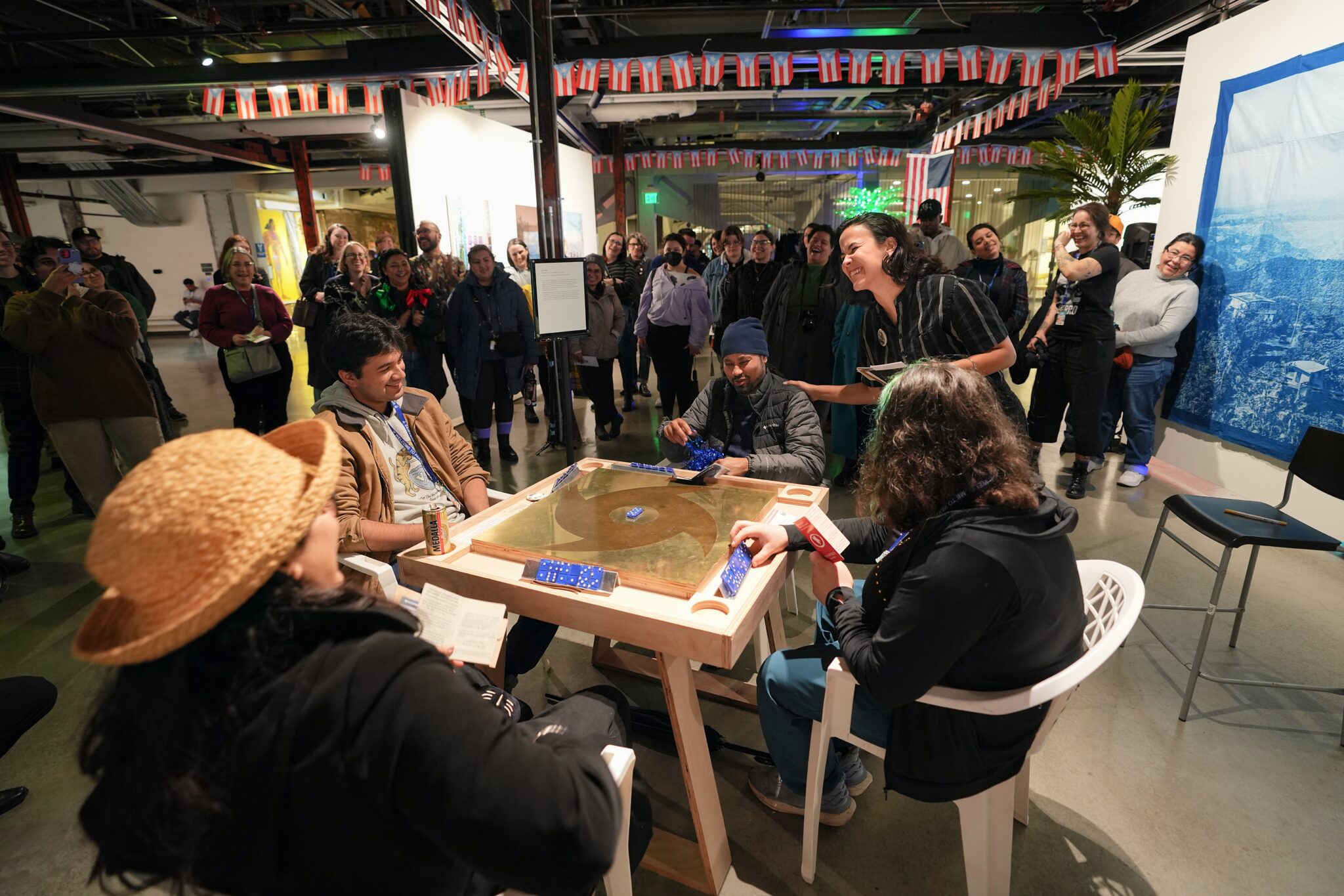 A group of four people sit around a square table in the middle of the gallery playing dominoes as a crowd looks on.