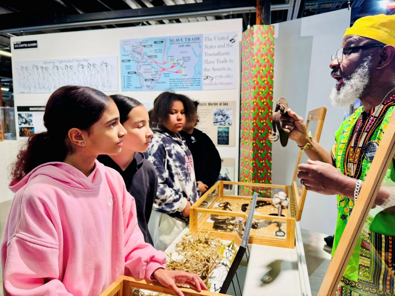An elder Black man shows historical artifacts to a group of four 5th grade students in a gallery.