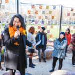 An Asian woman in a coat, orange scarf, and glasses speaks into a microphone amongst a dispersed crowd of listeners at the Seattle Center's Poetry Garden.