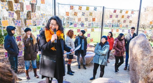 An Asian woman in a coat, orange scarf, and glasses speaks into a microphone amongst a dispersed crowd of listeners at the Seattle Center's Poetry Garden.