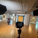 A behind-the-scenes shot of a professional film set inside an art gallery. A seated woman is framed by various artworks on the walls, including a large, colorful textile piece directly behind her.