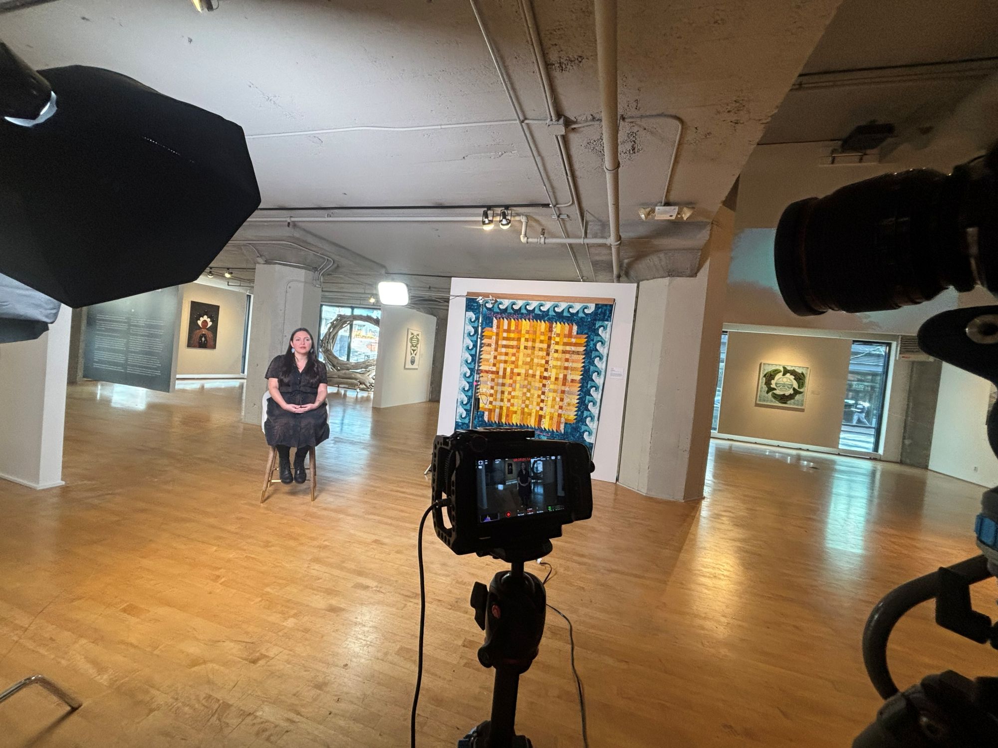 A behind-the-scenes shot of a professional film set inside an art gallery. A seated woman is framed by various artworks on the walls, including a large, colorful textile piece directly behind her.