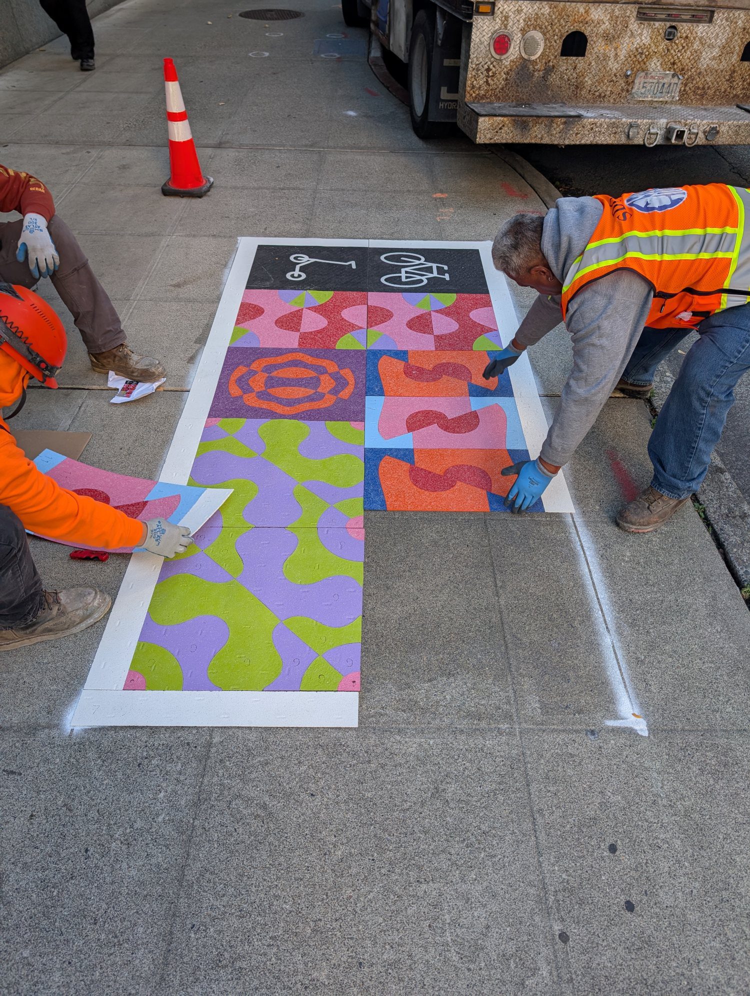 Construction workers place down colorful tiles on the sidewalk.
