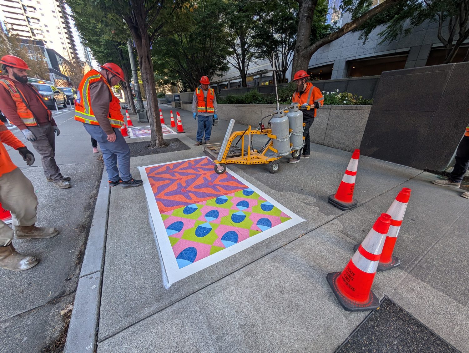 Construction workers with equipment inspect a colorful rectangular pattern on the sidewalk, surrounded by traffic cones.