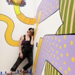 An artist wearing black overalls leans in a corner and smiles. The walls are painted with a colorful mural.