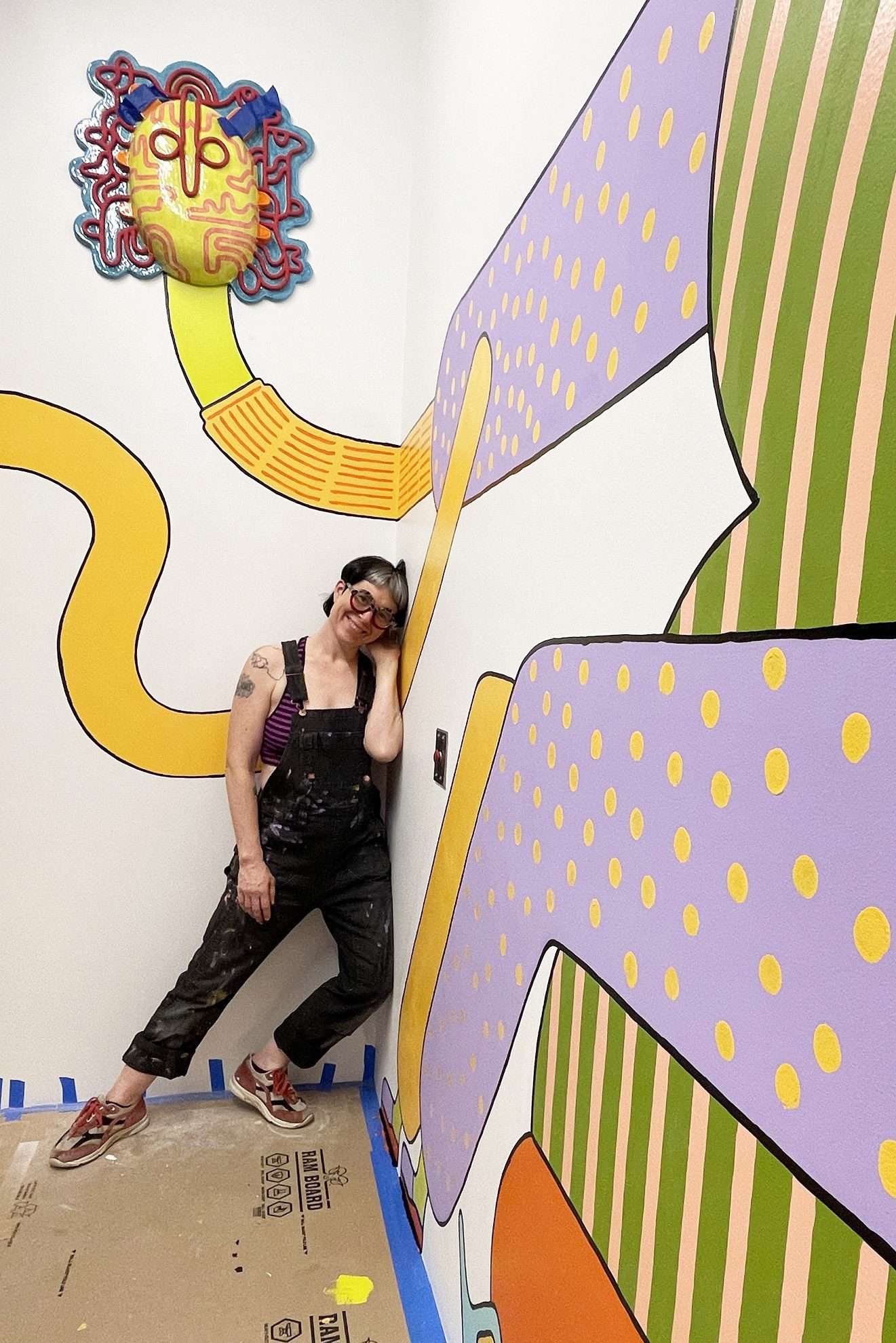 An artist wearing black overalls leans in a corner and smiles. The walls are painted with a colorful mural.