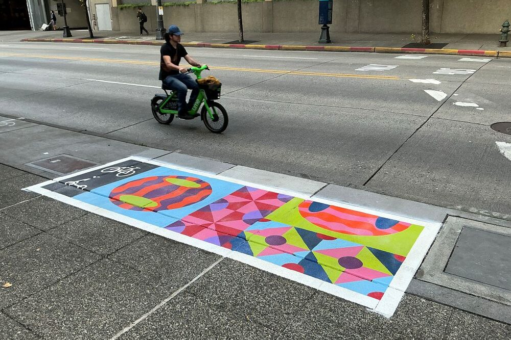 A colorful pattern painted in a rectangle on the sidewalk. A man on a Lime Bike drives by on the street.