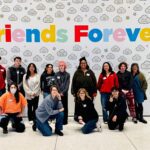A large group of teenagers posed together under a wall that has colorful lettering reading "Friends Forever"