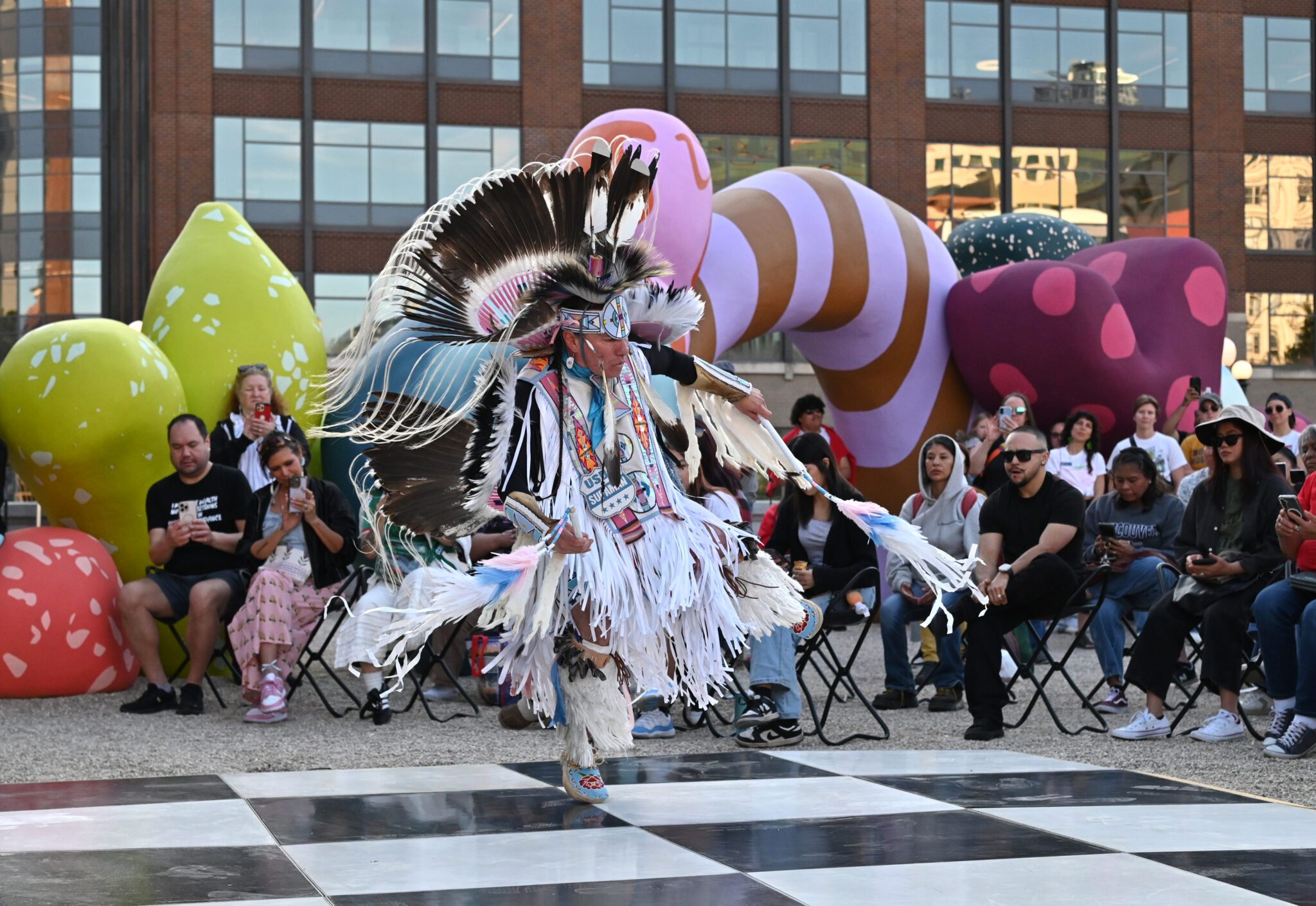 Supaman, an Indigenous artist, dresses in full regalia and is dancing on the plaza. An audience is seated behind him, with Clouds of Belonging behind them.