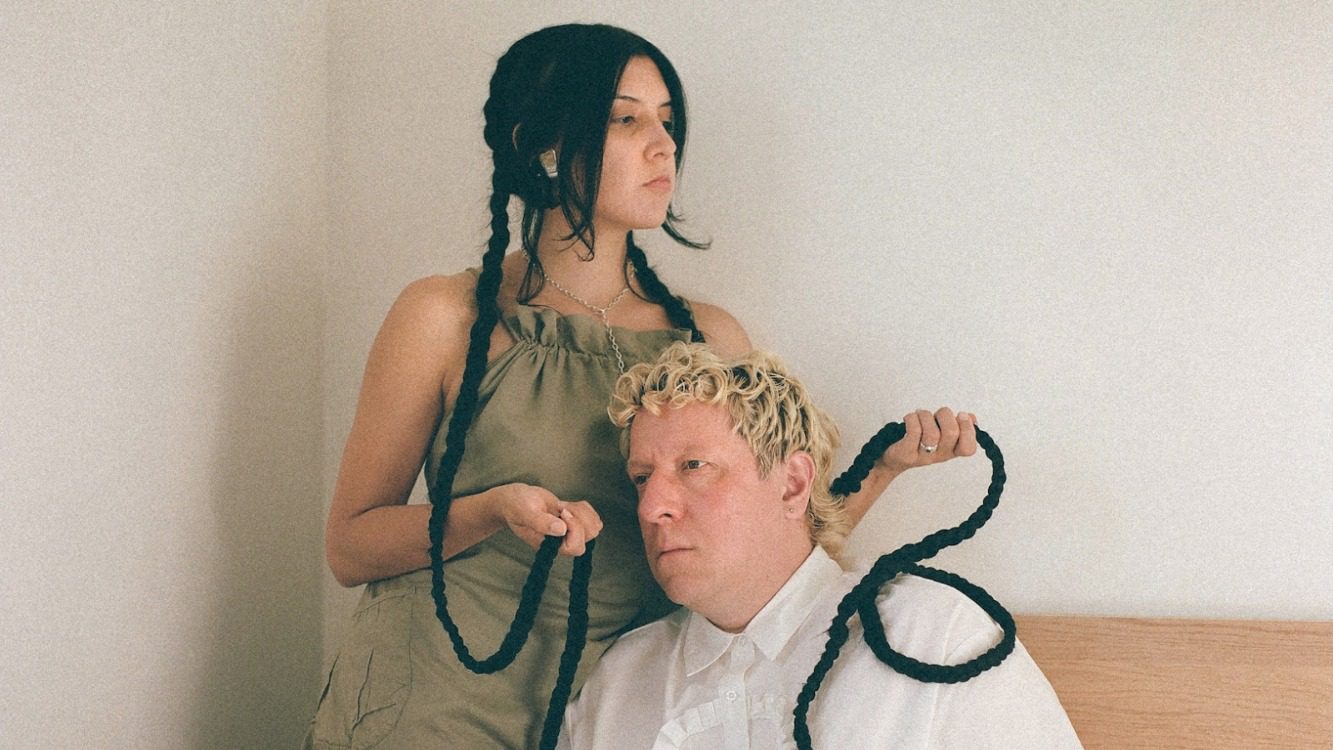 A woman with extremely long braids stands over a seated man and holder her braids over him.