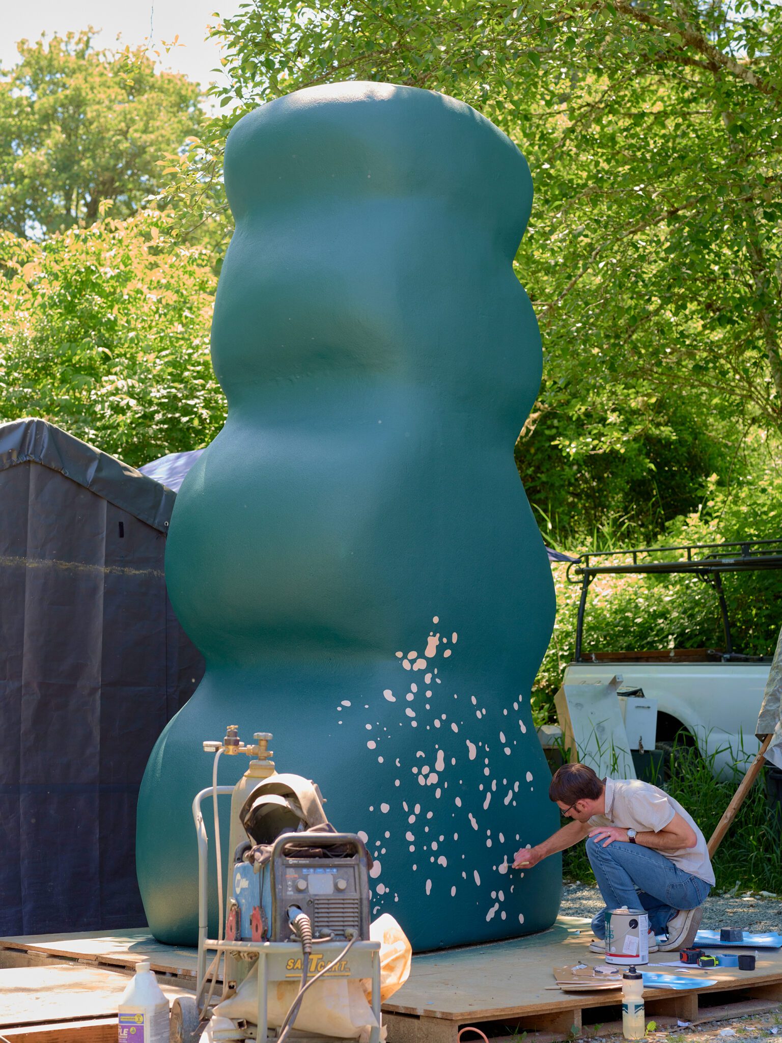 At the fabrication site; a person kneeling and painting pink paint on a tall teal curvy sculpture.