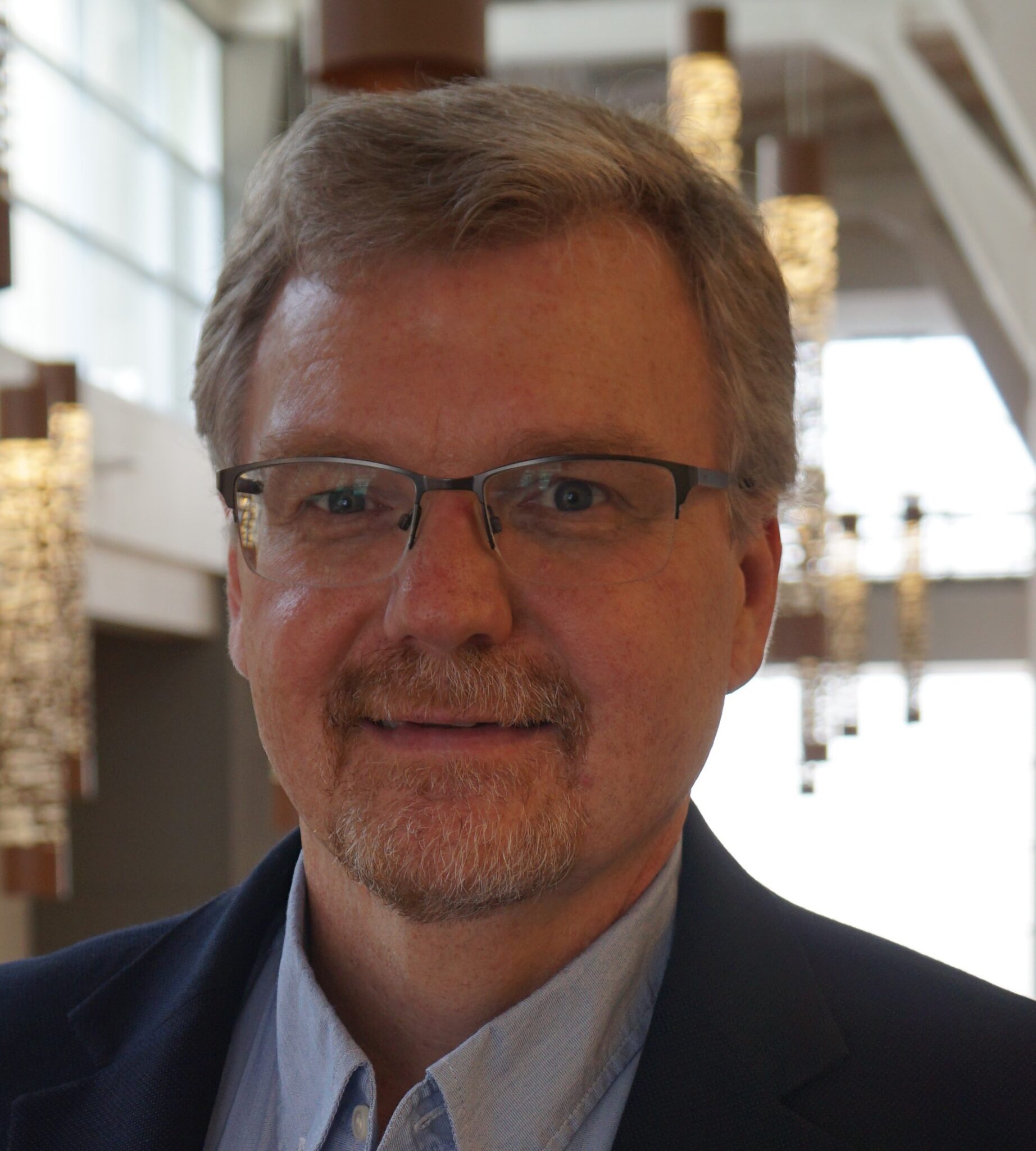 A headshot of a middle-aged white man in glasses wearing a navy suit.
