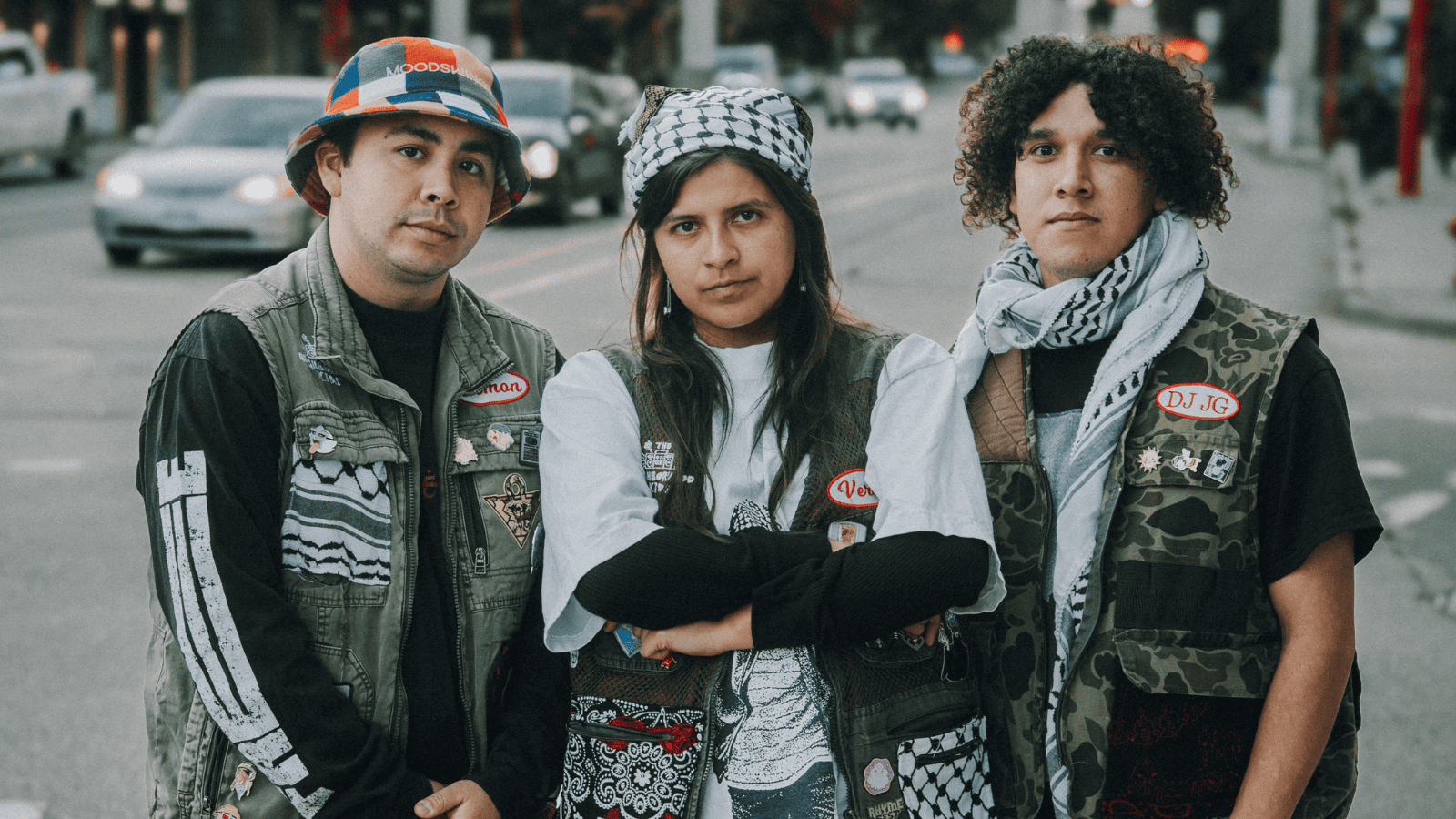 A group of three indigenous folks pose in camo vests wearing keffiyeh scarves.
