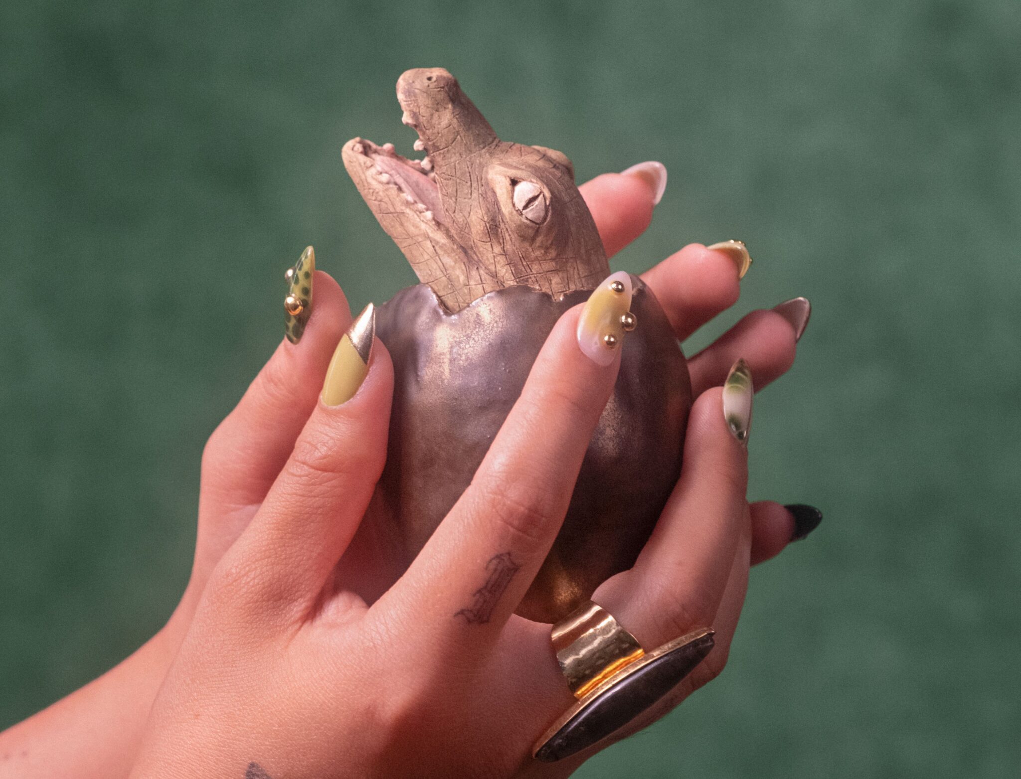 Two hands with intricate nail polish hold a ceramic egg with a baby crocodile emerging out of it