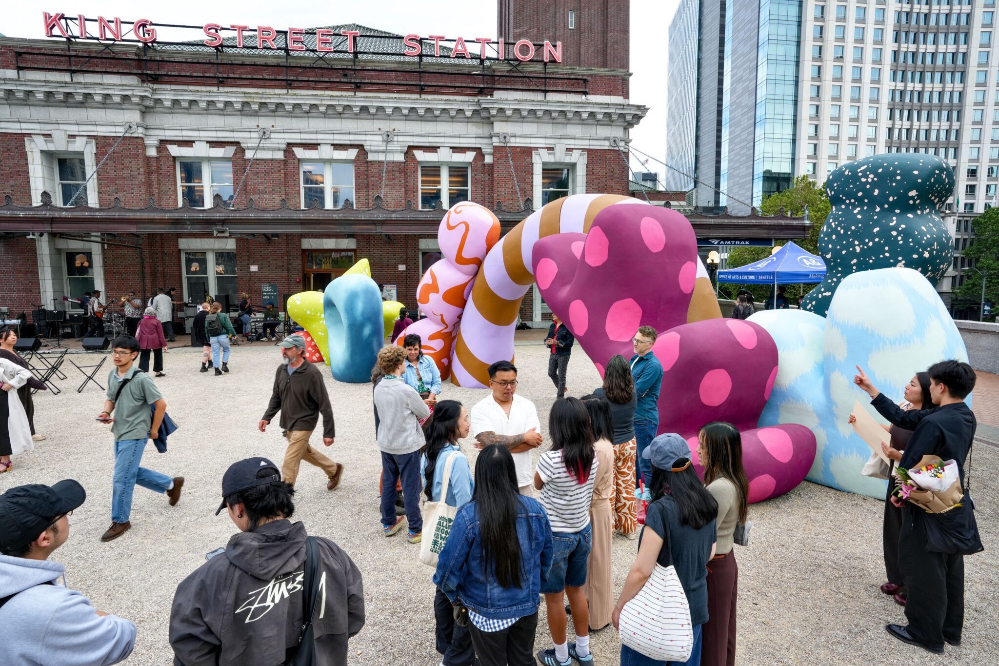 On the plaza of King Street Station sits a large colorful sculpture with an archway. People chat around it and walk through the arch.