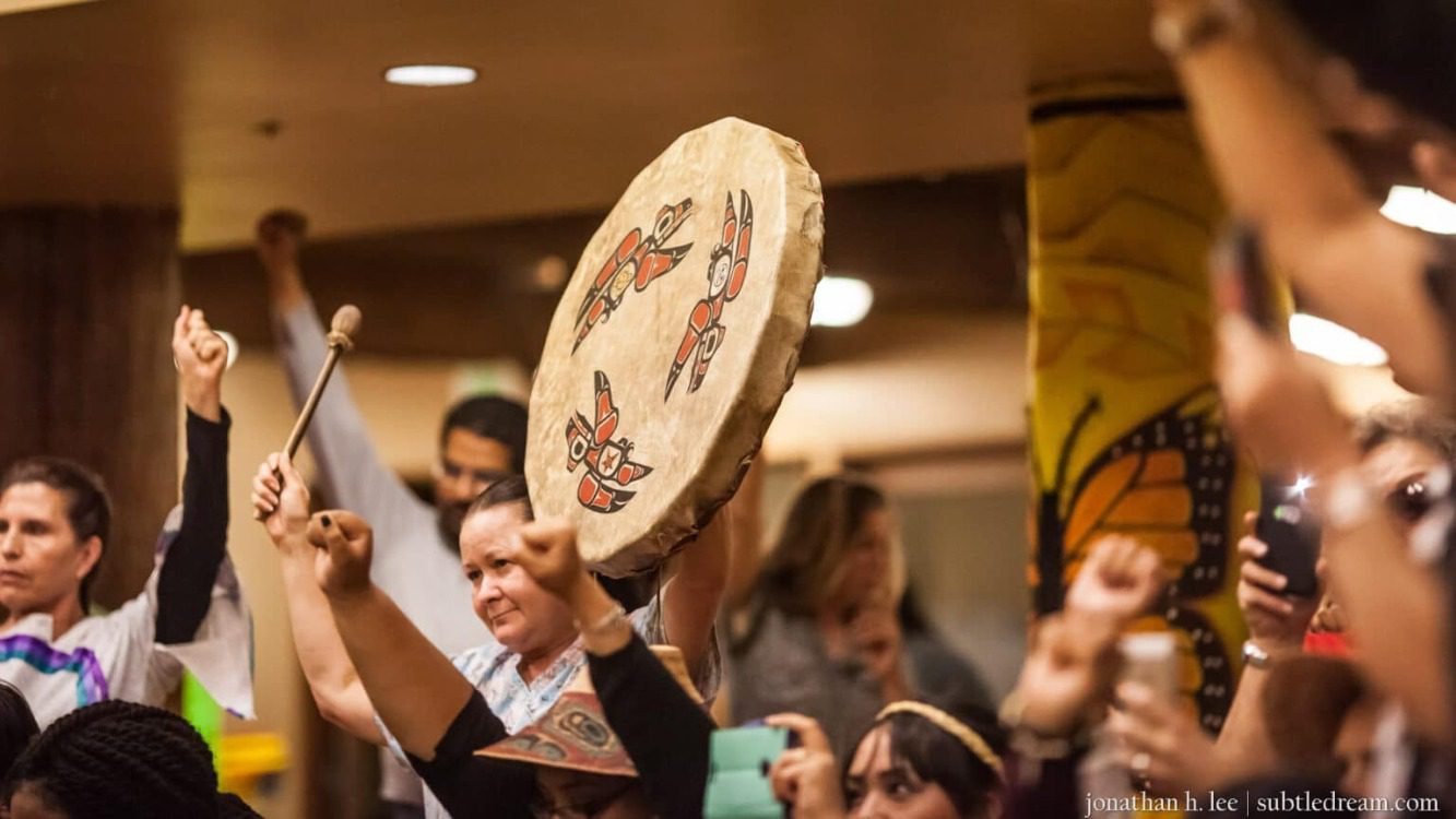 An Indigenous drum with paintings of birds is held up along with many people fists